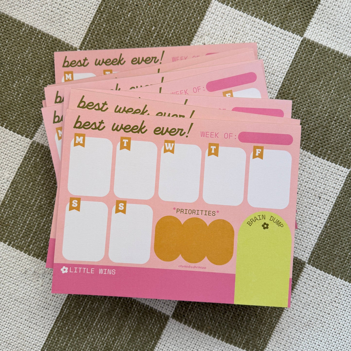 A stack of pink weekly planner notepads on a green and white checkered fabric background. The notepads feature sections for Monday through Friday, priority boxes, a brain dump area, and the text 'best week ever!' and 'WEEK OF:'.