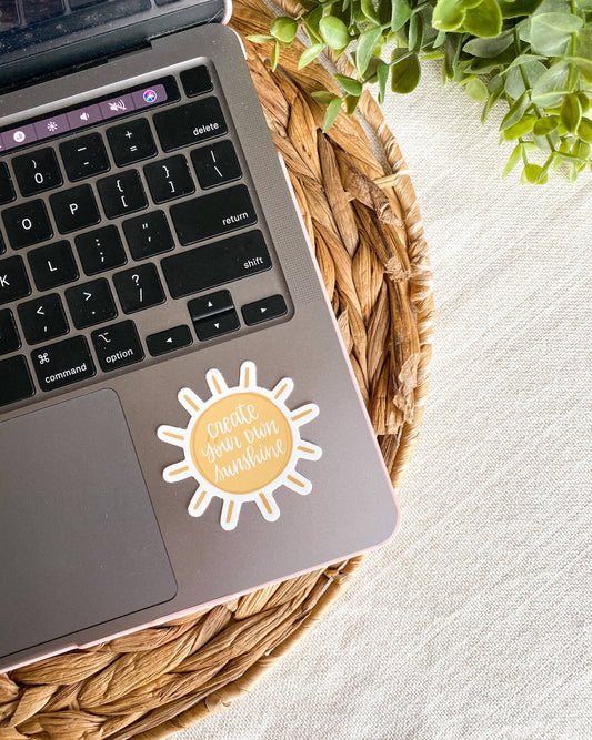 A die-cut sticker shaped like a sun with the phrase 'create your own sunshine' in gold script on a pale yellow center, shown applied to the palm rest of a laptop. The laptop sits on a woven straw placemat over a light fabric surface, with green leaves in the top-right corner.