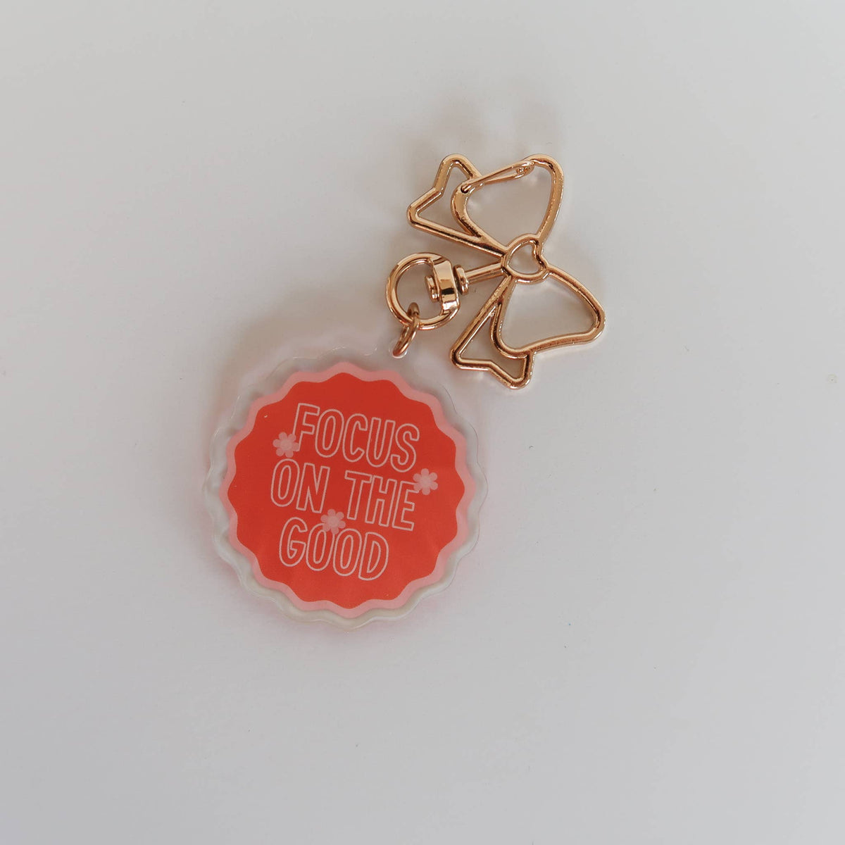 A small accessory charm with a scalloped orange-red circular face reading 'FOCUS ON THE GOOD' in white text, attached to a rose-gold metal bow and loop clasp, photographed on a plain light background.