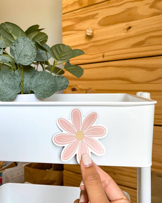 A hand with manicured nails is pressing a pink daisy-shaped Magnet onto the side of a white rolling storage cart. The background features a wooden wall paneling and a potted green plant in the upper left corner.