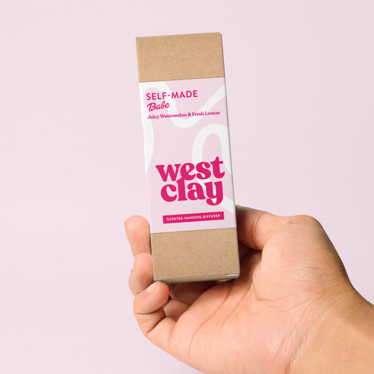 A hand holding a rectangular cardboard box for West Clay scented Air Freshener against a solid pale pink background. The box has a light pink label with Magenta text reading 'Self-Made Babe' and 'West Clay'.