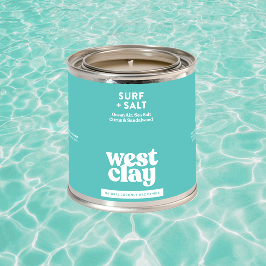 A tin candle labeled 'west clay' and 'SURF + SALT' sits centered on a turquoise water surface with rippled reflections. The product appears to be a wax candle in a metallic tin with a bright aqua label and white text.