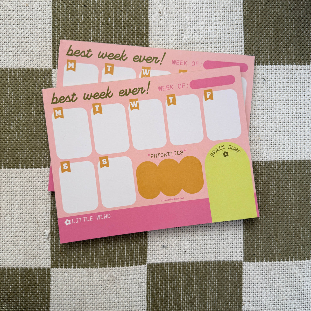 A pink weekly planner notepad with the phrase 'best week ever!' and a weekly grid, photographed on a brown and white checkered fabric background. The brand name 'LITTLE WINS' is visible in the bottom left corner.