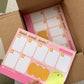 A person's hand peeling back a yellow sticky note from a pink weekly planner pad, which is sitting inside an open brown cardboard box on a green and white checkered floor.