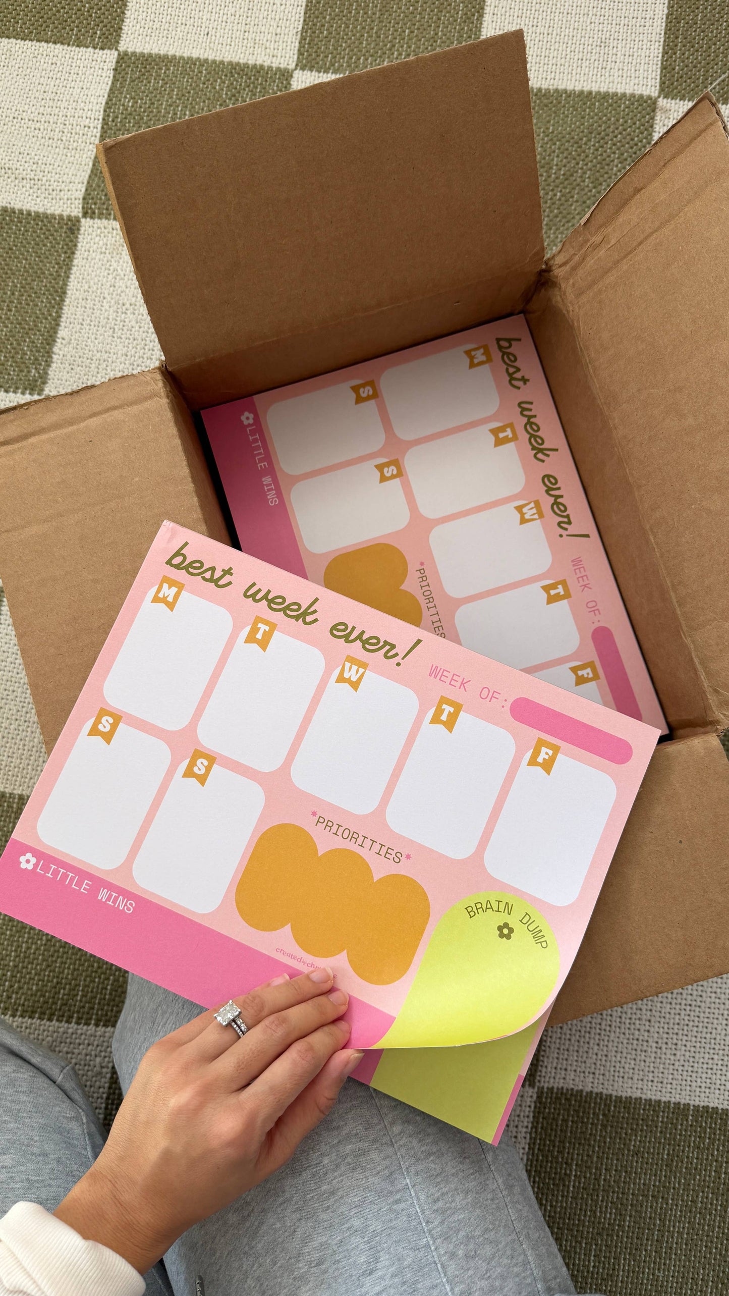 A person's hand peeling back a yellow sticky note from a pink weekly planner pad, which is sitting inside an open brown cardboard box on a green and white checkered floor.