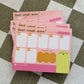 A stack of pink weekly planner notepads on a green and white checkered fabric background. The notepads feature sections for Monday through Friday, priority boxes, a brain dump area, and the text 'best week ever!' and 'WEEK OF:'.