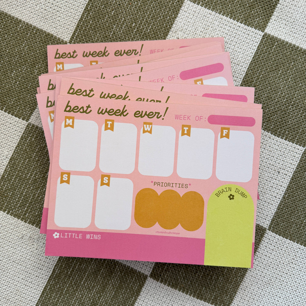 A stack of pink weekly planner notepads on a green and white checkered fabric background. The notepads feature sections for Monday through Friday, priority boxes, a brain dump area, and the text 'best week ever!' and 'WEEK OF:'.