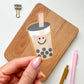 A hand holds a die-cut sticker of a smiling boba tea cup in front of a wooden board, with a metal binder clip and pink and gold pens on a white surface.