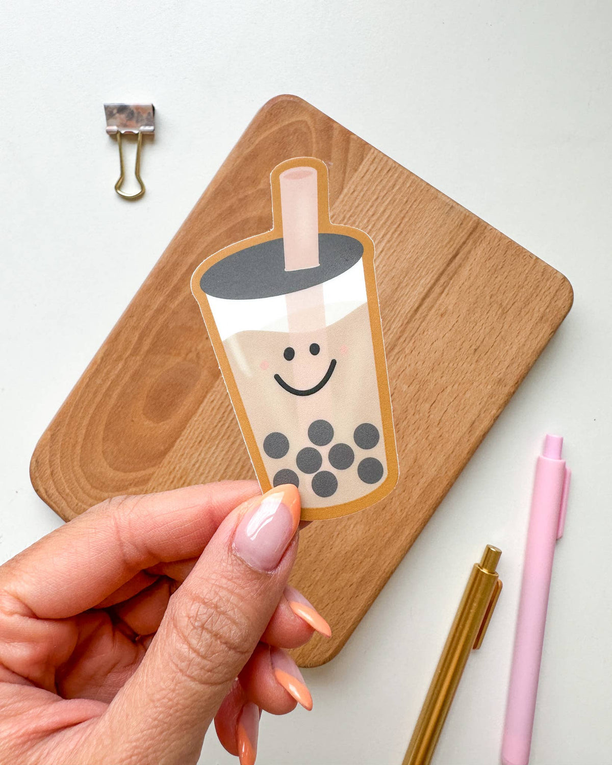 A hand holds a die-cut sticker of a smiling boba tea cup in front of a wooden board, with a metal binder clip and pink and gold pens on a white surface.