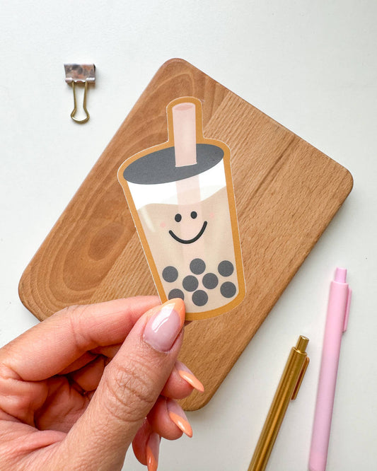 A hand holds a die-cut sticker of a smiling boba tea cup in front of a wooden board, with a metal binder clip and pink and gold pens on a white surface.