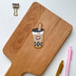 A die-cut sticker of a smiling boba tea cup with black pearls, placed on a wooden cutting board. The board is styled on a white background with a gold binder clip, a pink pen, and a gold pen.