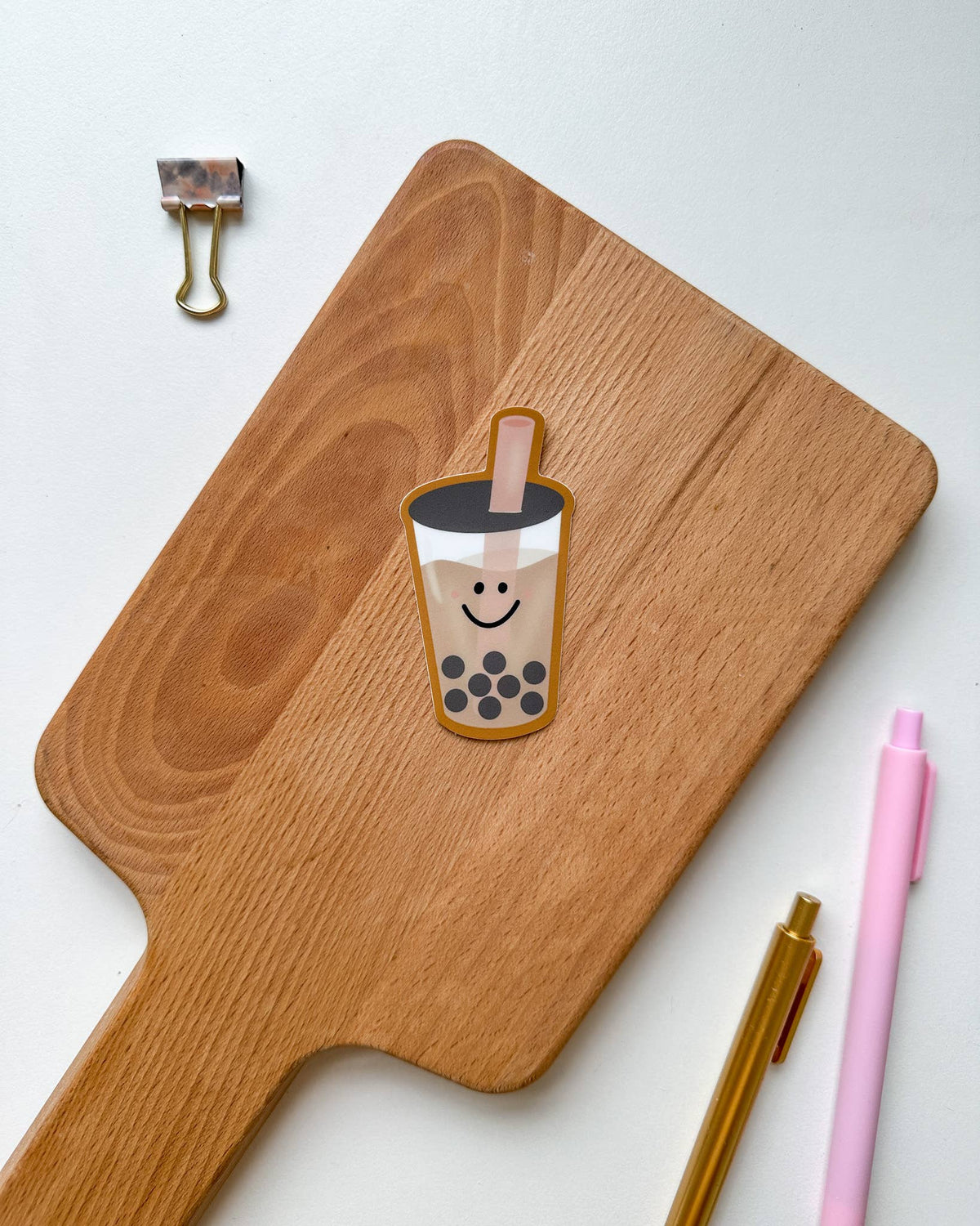 A die-cut sticker of a smiling boba tea cup with black pearls, placed on a wooden cutting board. The board is styled on a white background with a gold binder clip, a pink pen, and a gold pen.