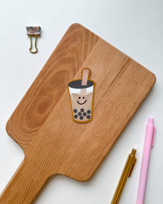 A die-cut sticker of a smiling boba tea cup with black pearls, placed on a wooden cutting board. The board is styled on a white background with a gold binder clip, a pink pen, and a gold pen.