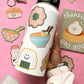 A hand holds a white insulated water bottle decorated with cute sushi and ramen illustrations, set against a pink background with matching sushi stickers.