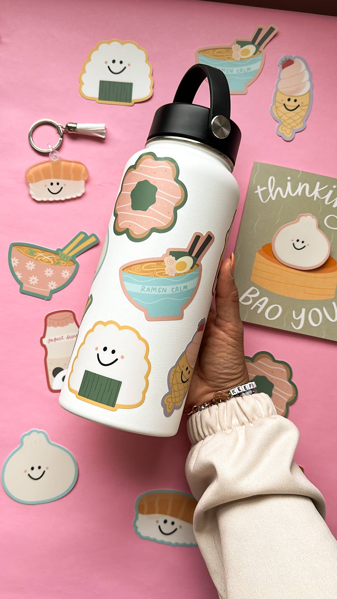 A hand holds a white insulated water bottle decorated with cute sushi and ramen illustrations, set against a pink background with matching sushi stickers.