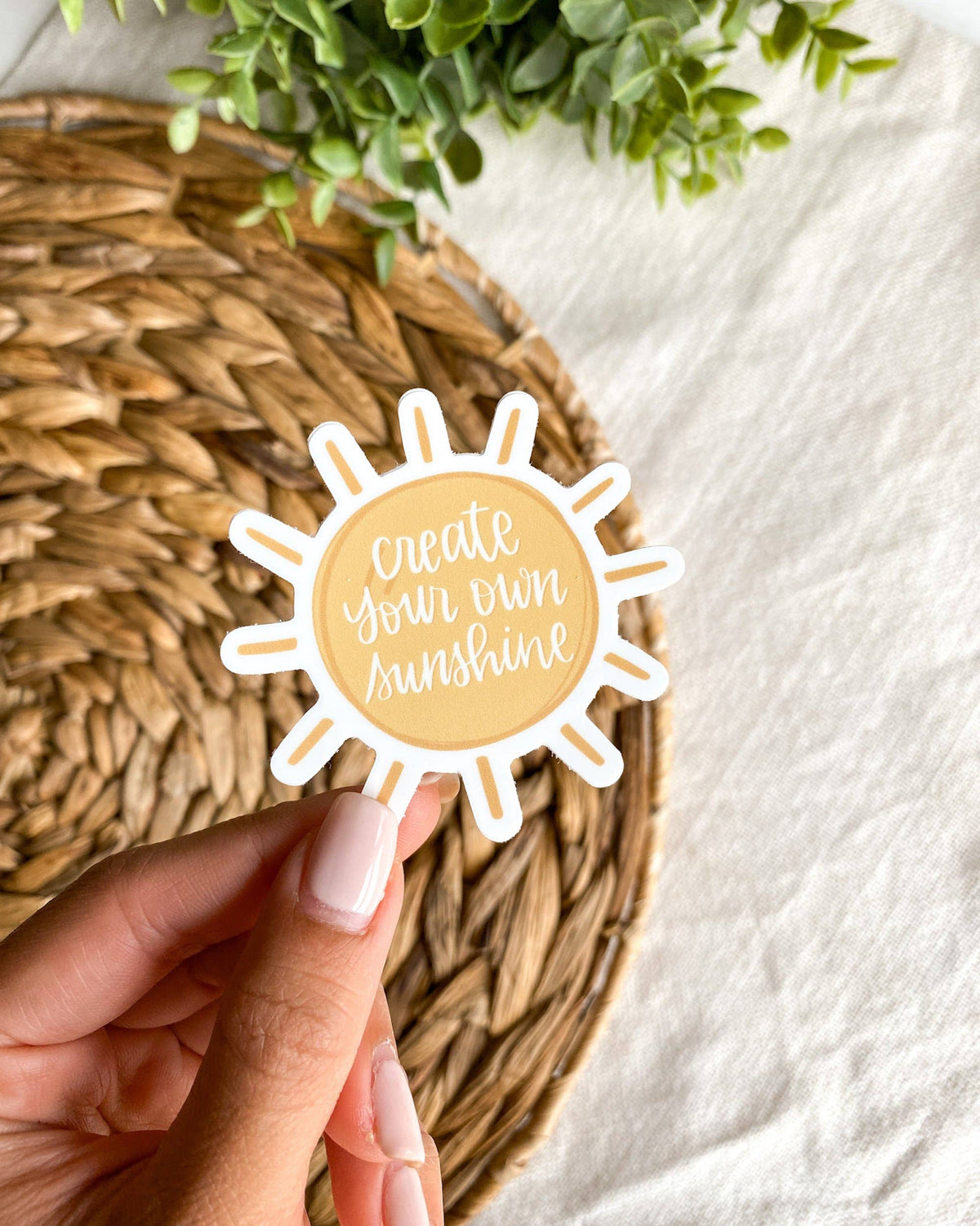 A hand with light pink manicured nails holds a die-cut sticker shaped like a sun with the text 'create your own sunshine' in white script on a pale yellow center. The sticker is positioned over a round woven straw placemat on a white fabric surface, with green leaves visible in the top corner.