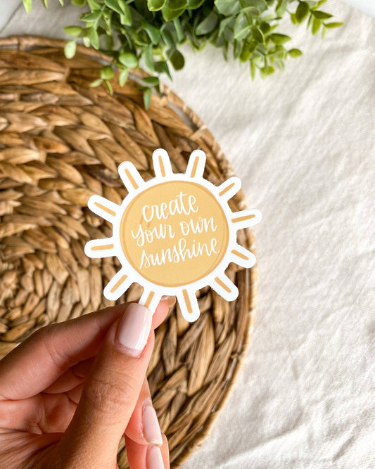 A hand with light pink manicured nails holds a die-cut sticker shaped like a sun with the text 'create your own sunshine' in white script on a pale yellow center. The sticker is positioned over a round woven straw placemat on a white fabric surface, with green leaves visible in the top corner.