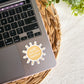 A die-cut sticker shaped like a sun with the phrase 'create your own sunshine' in gold script on a pale yellow center, shown applied to the palm rest of a laptop. The laptop sits on a woven straw placemat over a light fabric surface, with green leaves in the top-right corner.