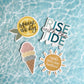 Four die-cut sticker designs arranged on a blue water surface with rippled reflections. The stickers include a lemon with the phrase 'squeeze the day,' a multicolor ice cream cone, a white and teal block-letter phrase 'RISE with the TIDE,' and a sun with the phrase 'create your own sunshine.