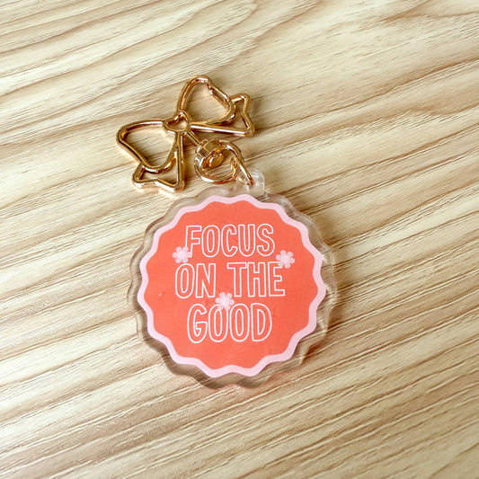 A scalloped acrylic keychain charm with a gold metal bow clasp, featuring a coral pink circular design with the text 'FOCUS ON THE GOOD' in white, resting on a light wood grain surface.