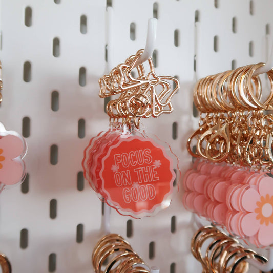 A close-up of a pink scalloped acrylic charm with the words 'FOCUS ON THE GOOD' in white, attached to a rose gold metal keychain, hanging on a white pegboard display.