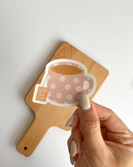 A hand with beige manicured nails holds a die-cut sticker of a pink floral teacup with the words 'still the tea' next to a small tea bag icon. The sticker is positioned in front of a light wood cutting board on a clean white background.