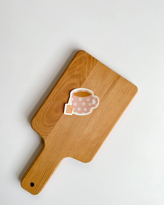 A small die-cut sticker shaped like a pink teacup with white floral patterns and a tea bag tag, placed on a light wood cutting board against a white background.