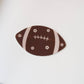 A football-shaped sticker in dark brown with white stripes, laces, and small white flower motifs, shown on a plain white background.