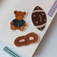 Three cute die-cut stickers displayed on a white notebook page. The stickers feature a brown teddy bear wearing a navy blue jersey, a dark brown football with cream stripes, and a pretzel with a pink filling.