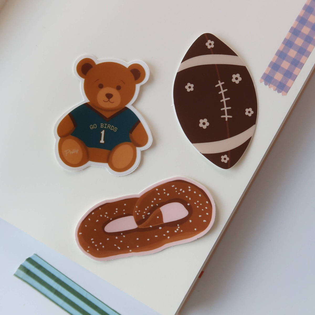 Three cute die-cut stickers displayed on a white notebook page. The stickers feature a brown teddy bear wearing a navy blue jersey, a dark brown football with cream stripes, and a pretzel with a pink filling.