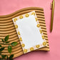 A flat-lay of a notepad with a white center and a border of yellow smiley faces on a pink background, styled on a wavy wooden tray with a gold pen and green leaves.