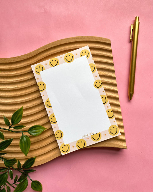 A flat-lay of a notepad with a white center and a border of yellow smiley faces on a pink background, styled on a wavy wooden tray with a gold pen and green leaves.