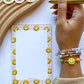 A hand holding a gold pen next to a notepad with a smiley face border, featuring colorful smiley face stickers and a wooden tray on a white background.