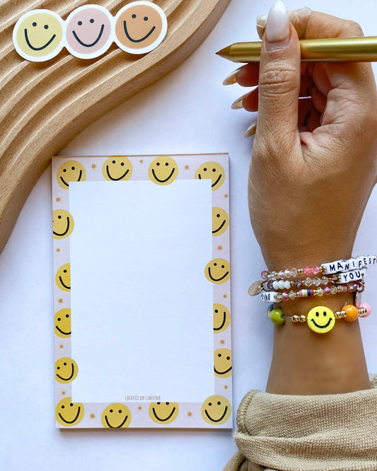 A hand holding a gold pen next to a notepad with a smiley face border, featuring colorful smiley face stickers and a wooden tray on a white background.