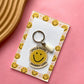 A yellow smiley face acrylic keychain with the text 'SMILE MORE WORRY LESS' and a small white tassel, displayed on a printed backing card with smiley faces, set on a pink background with a wooden tray corner visible.