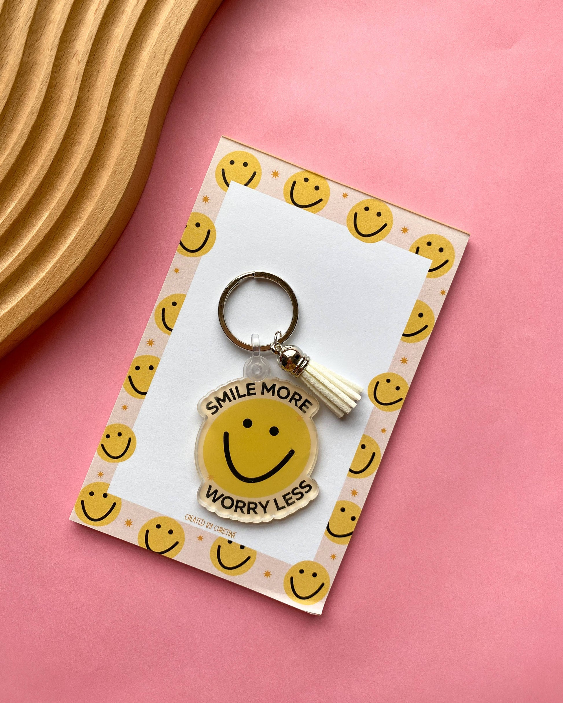 A yellow smiley face acrylic keychain with the text 'SMILE MORE WORRY LESS' and a small white tassel, displayed on a printed backing card with smiley faces, set on a pink background with a wooden tray corner visible.