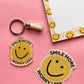 A yellow smiley face keychain with a tassel and matching die-cut sticker on a pink background, next to a matching yellow smiley face notepad.