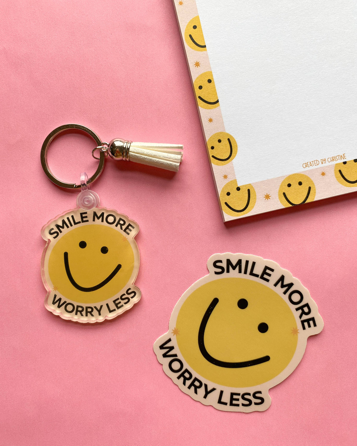 A yellow smiley face keychain with a tassel and matching die-cut sticker on a pink background, next to a matching yellow smiley face notepad.