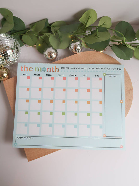 A monthly calendar notepad with a light blue border and colorful day markers, styled on a wooden board with green leaves, a disco ball, and silver ornaments.