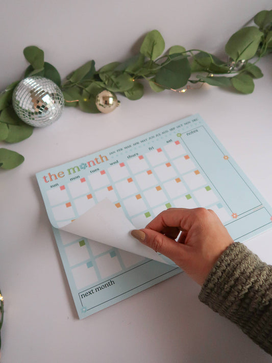 A hand peeling back a page from a pastel blue monthly planner pad decorated with green leaves and holiday ornaments on a white surface.