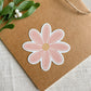 A die-cut Magnet shaped like a daisy flower with pink petals, a white border, and a beige center, placed on a brown kraft paper envelope with green leaves in the corner.