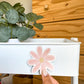 A hand with manicured nails is pressing a pink daisy-shaped Magnet onto the side of a white rolling storage cart. The background features a wooden wall paneling and a potted green plant in the upper left corner.