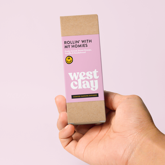 A hand holding a rectangular cardboard box for West Clay scented wax melts against a solid pale pink background. The box has a light pink label with white text reading 'ROLLIN' WITH MY HOMIES' and 'west clay', plus a small yellow smiley face logo.