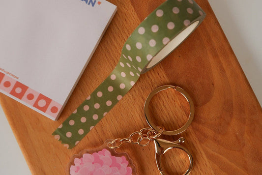 A styled flat-lay of a green and pink polka dot washi tape roll on a wooden surface, alongside a white notepad and a rose gold keychain with a pink flower charm.