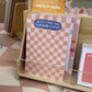 A wooden display stand holding pastel stationery items, including a pink-and-white checkered notepad with a blue sticker that reads 'WRITE IT DOWN NOW. OVERTHINK IT LATER!' and other cards and pads in soft peach, pink, yellow, and green tones.