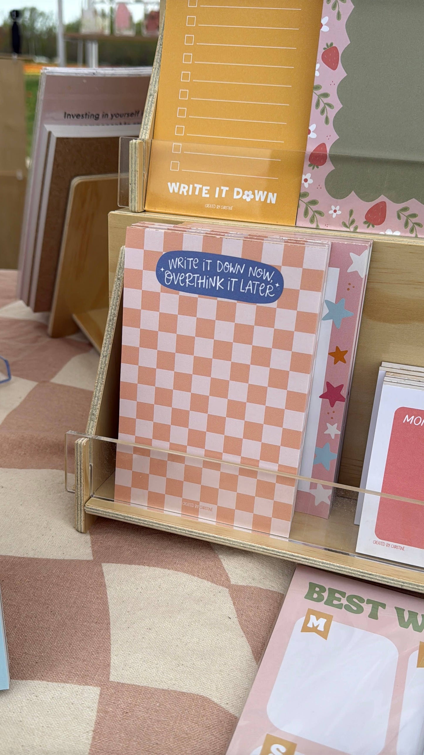 A wooden display stand holding pastel stationery items, including a pink-and-white checkered notepad with a blue sticker that reads 'WRITE IT DOWN NOW. OVERTHINK IT LATER!' and other cards and pads in soft peach, pink, yellow, and green tones.